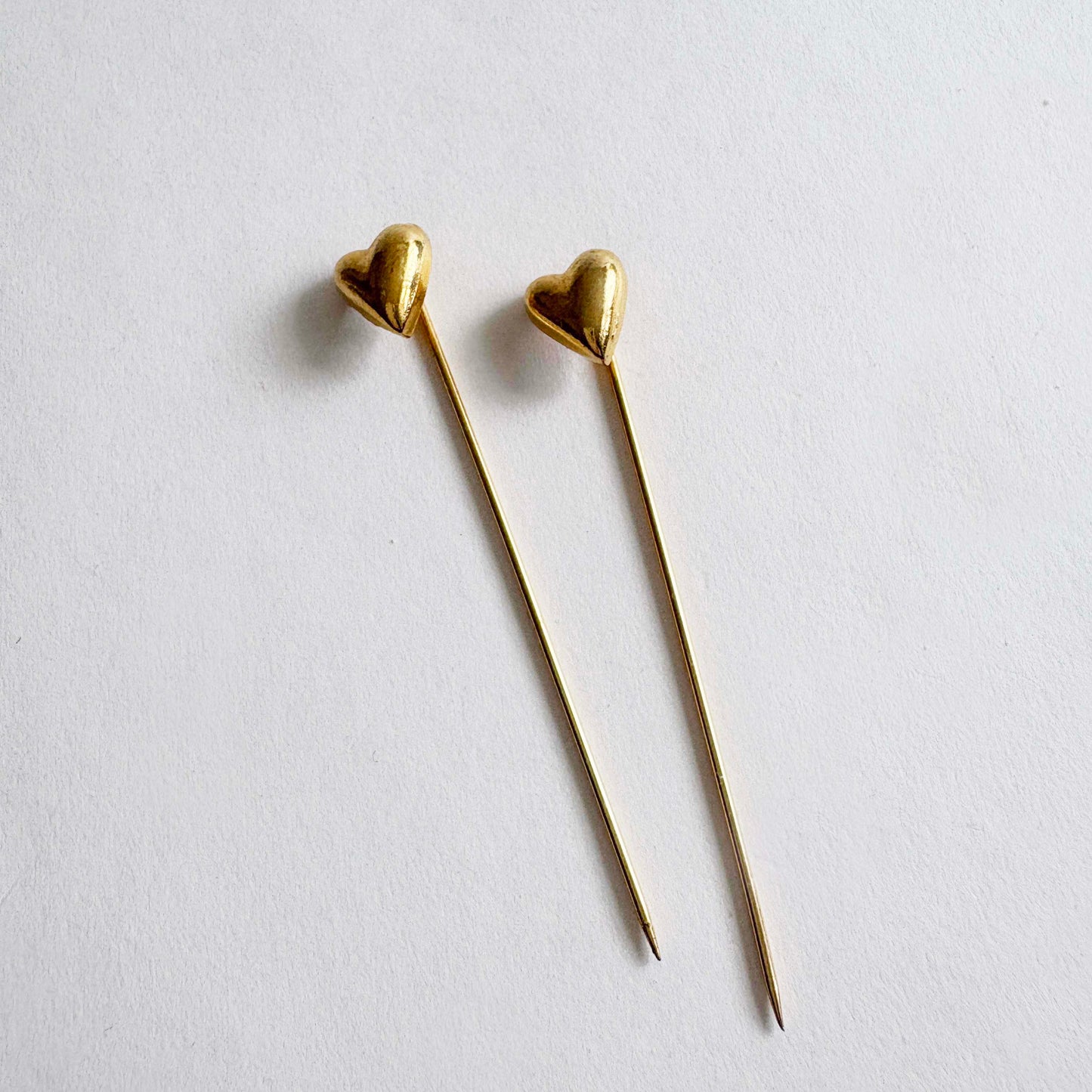 Pair of vintage gold-tone heart stick pins with long slender needles on white background