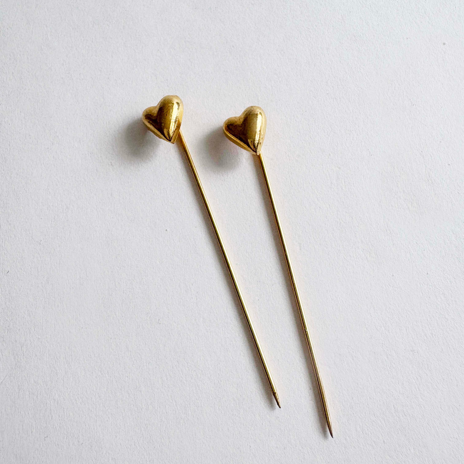 Pair of vintage gold-tone heart stick pins with long slender needles on white background