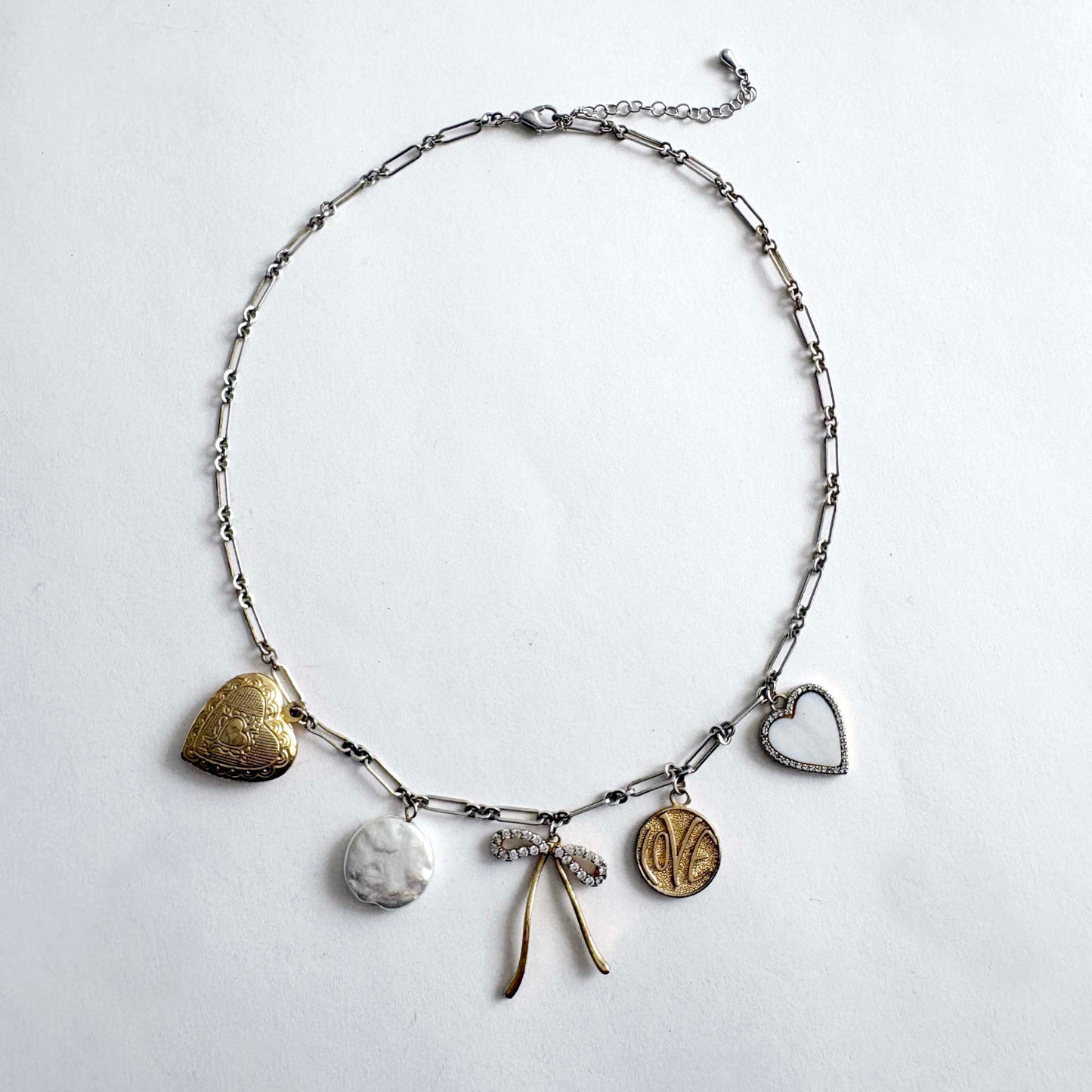 Silver chain charm necklace with gold heart locket, crystal bow, love coin and mother of pearl heart charms on white background