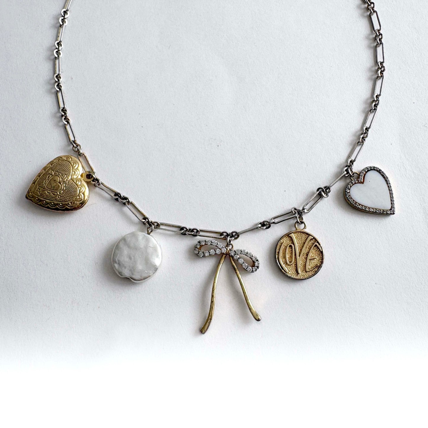 Silver chain charm necklace with gold heart locket, crystal bow, love coin and mother of pearl heart charms on white background