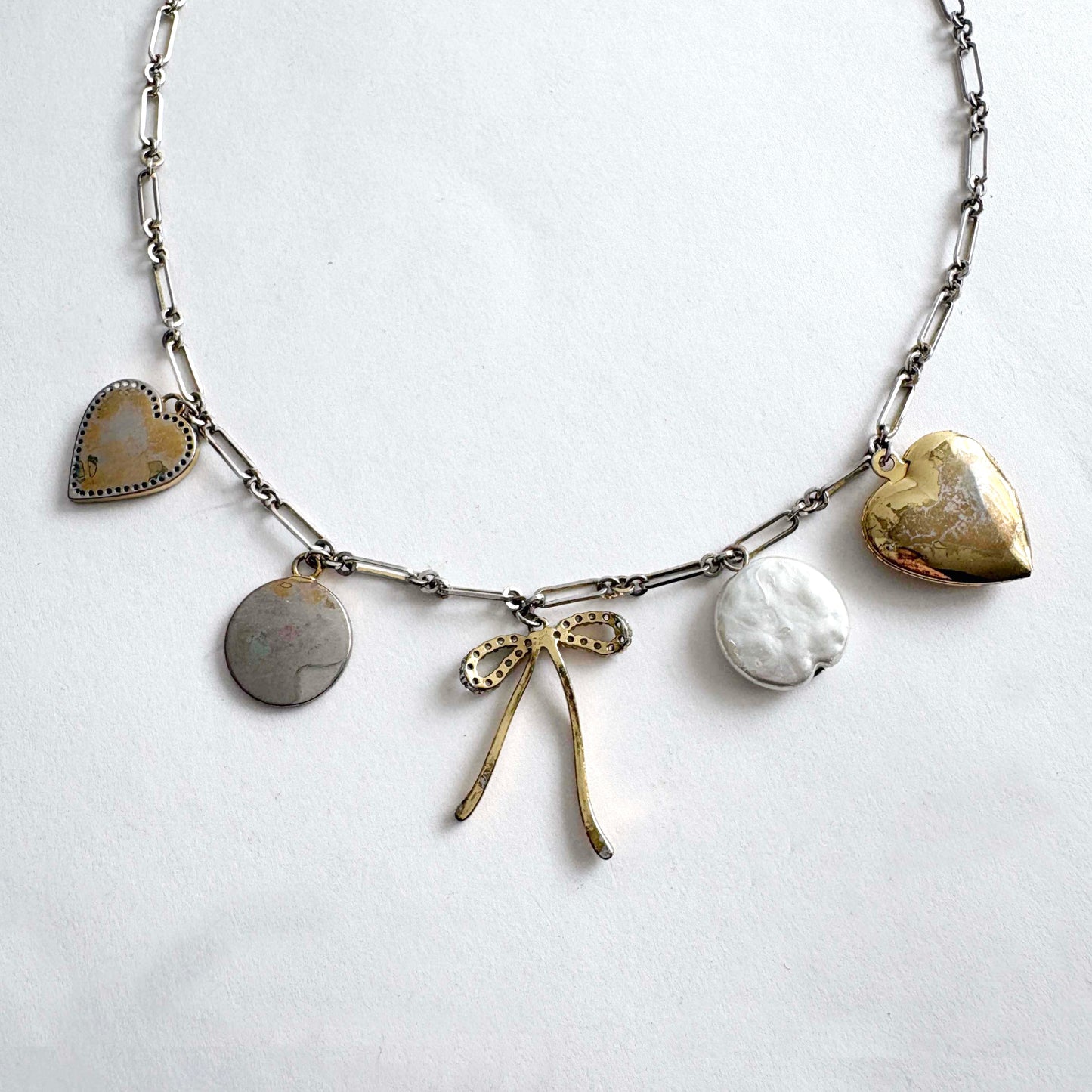 Silver chain charm necklace (back) with gold heart locket, crystal bow, love coin and mother of pearl heart charms on white background