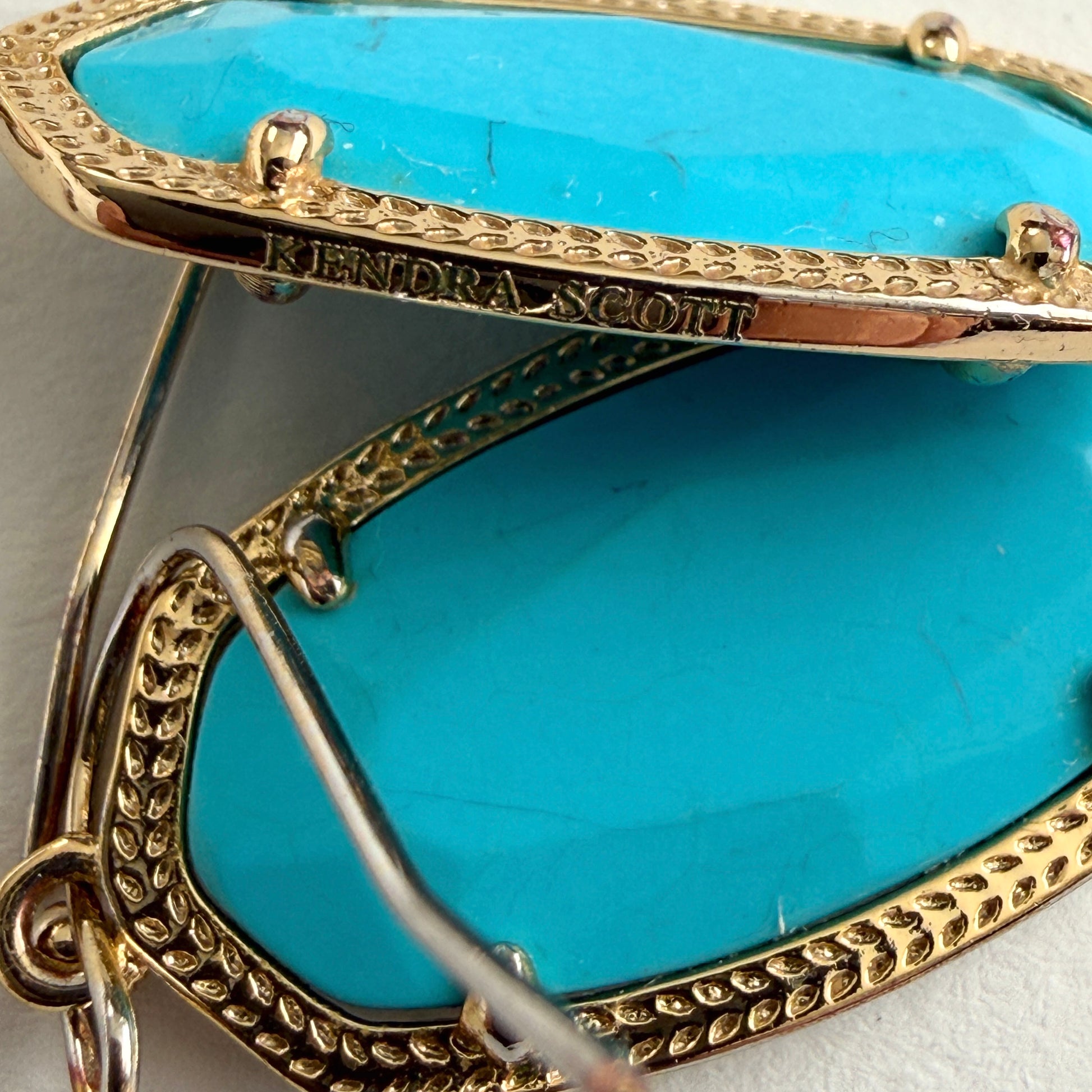 Close-up of a turquoise earring with gold accents and 'Kendra Scott' branding.